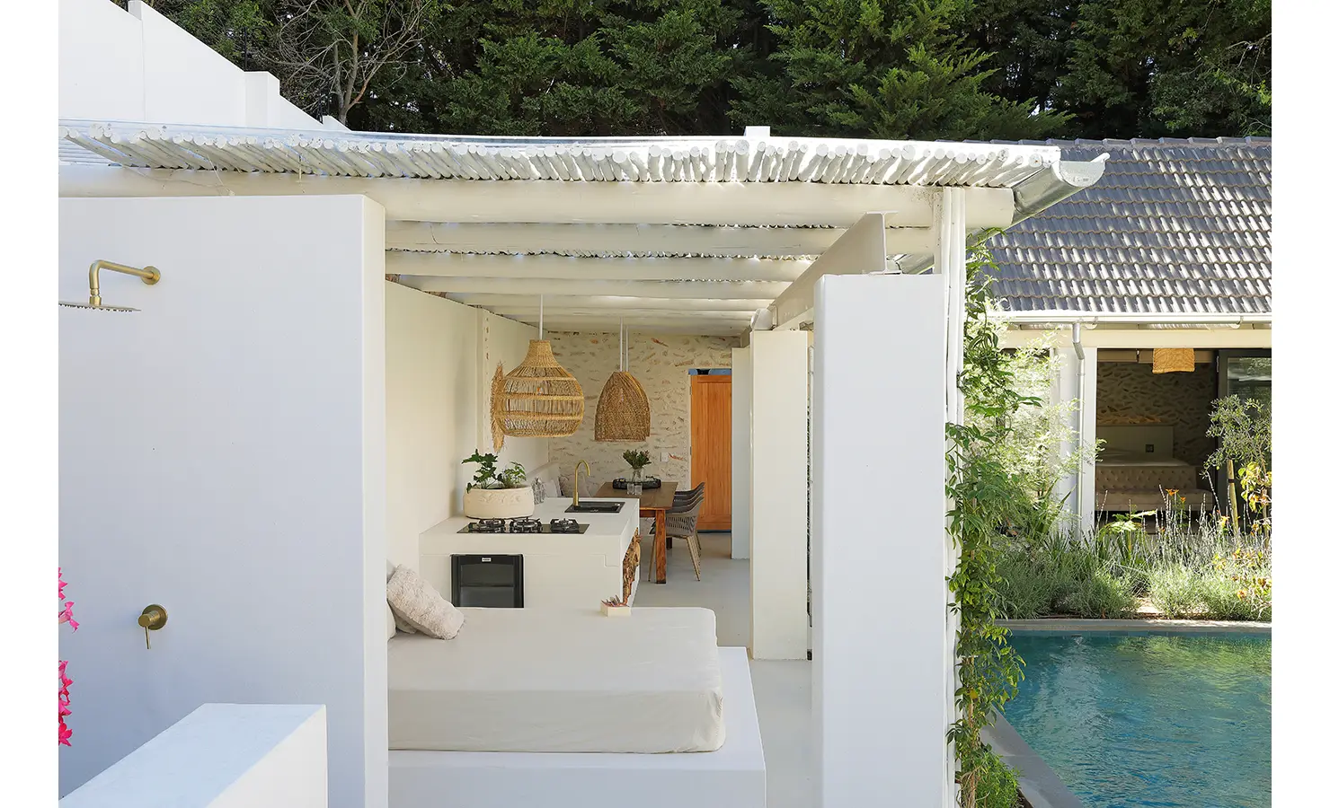 Perfect Hideaways: A breezy outdoor kitchen and lounge area under a slatted roof, featuring woven pendant lights, pale stone walls, and views of the glistening pool. Bel Ombre Hills, Constantia.