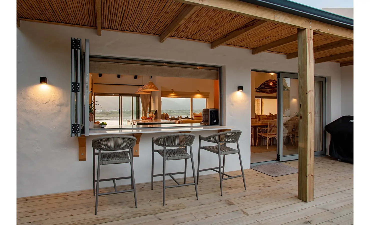 Perfect Hideaways: An inviting outdoor bar with high stools and a hatch-style window looks into the kitchen and living areas, blending indoor warmth with the outdoor deck. River Run, Plettenberg Bay.