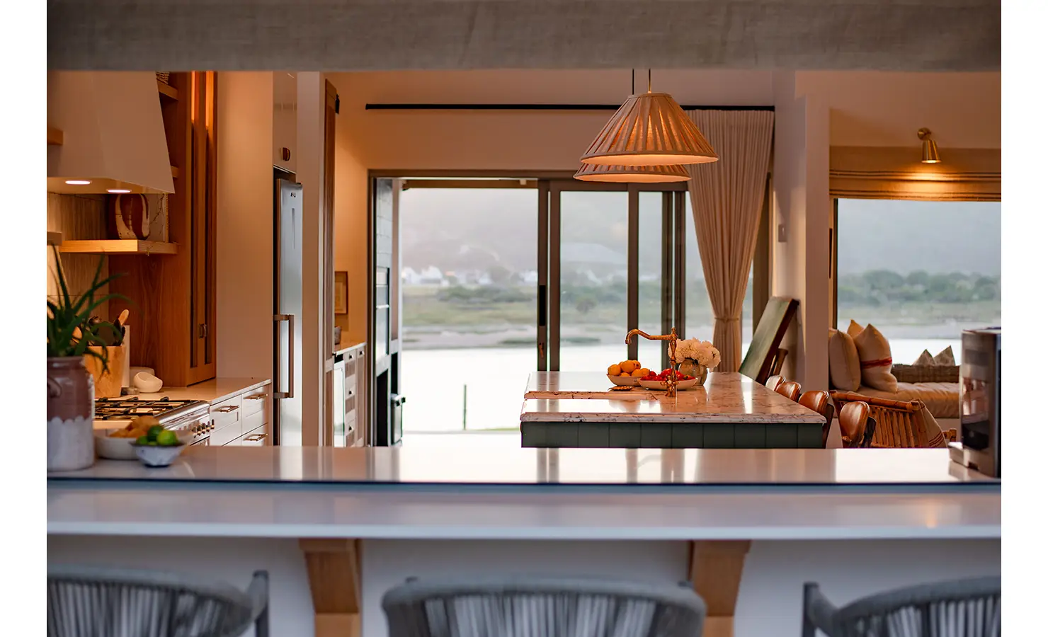 Perfect Hideaways: A view from the outdoor bar peers through to the kitchen island, cosy lounge, and river beyond, capturing the seamless indoor-outdoor connection. River Run, Plettenberg Bay.