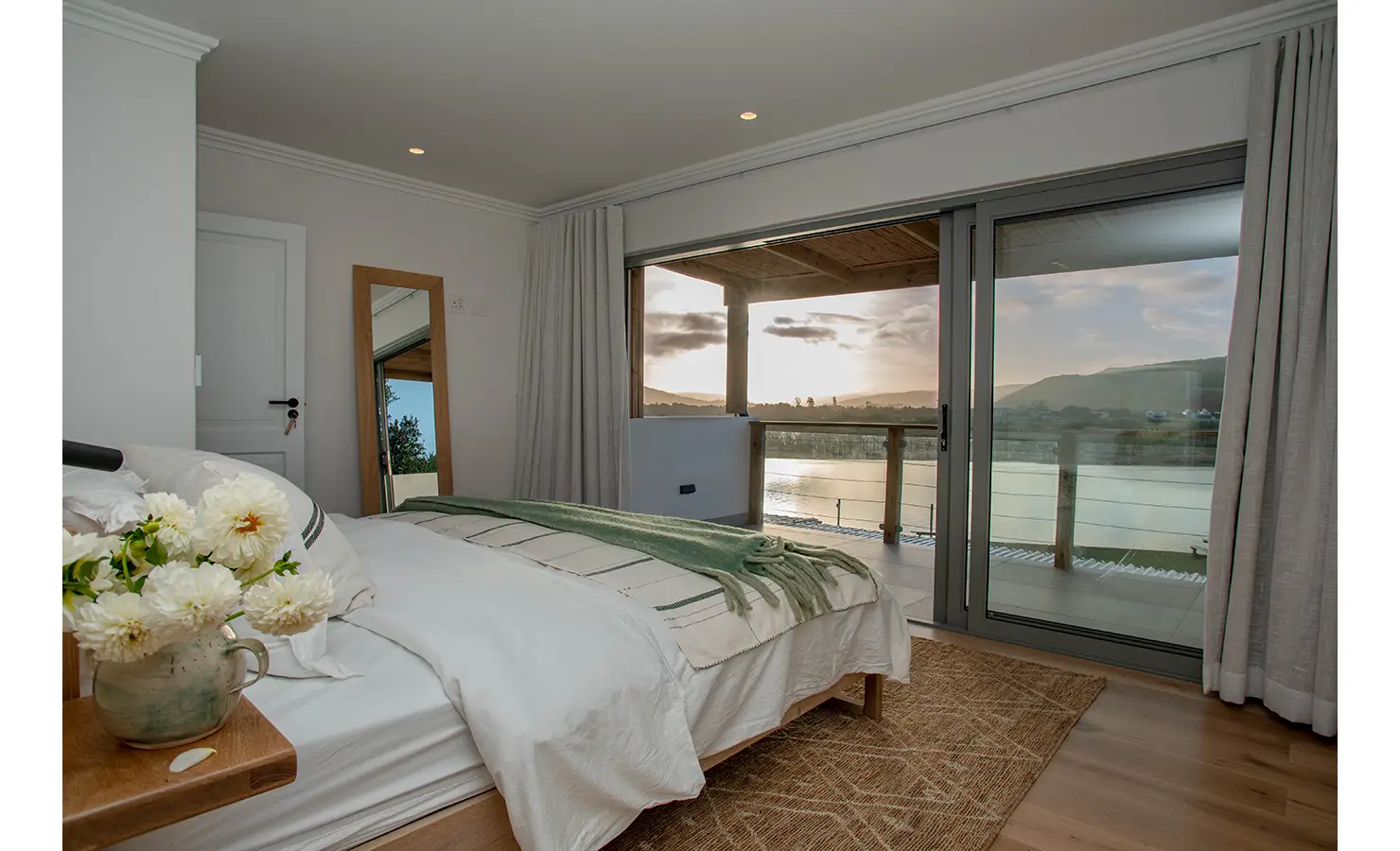 Perfect Hideaways: A serene bedroom with neutral tones, a green throw, and fresh flowers opens onto a covered balcony overlooking the river at dusk. River Run, Plettenberg Bay.