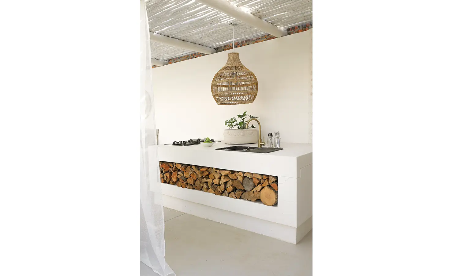 Perfect Hideaways: A sleek white outdoor kitchen with wood storage, soft lighting, and botanical touches beneath a stylish woven light fixture. Bel Ombre Hills, Constantia.