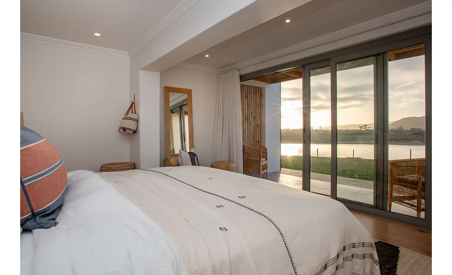 Perfect Hideaways: A calming bedroom with natural textures, a striped blanket, and soft lighting opens to river views through floor-to-ceiling sliding doors. River Run, Plettenberg Bay.