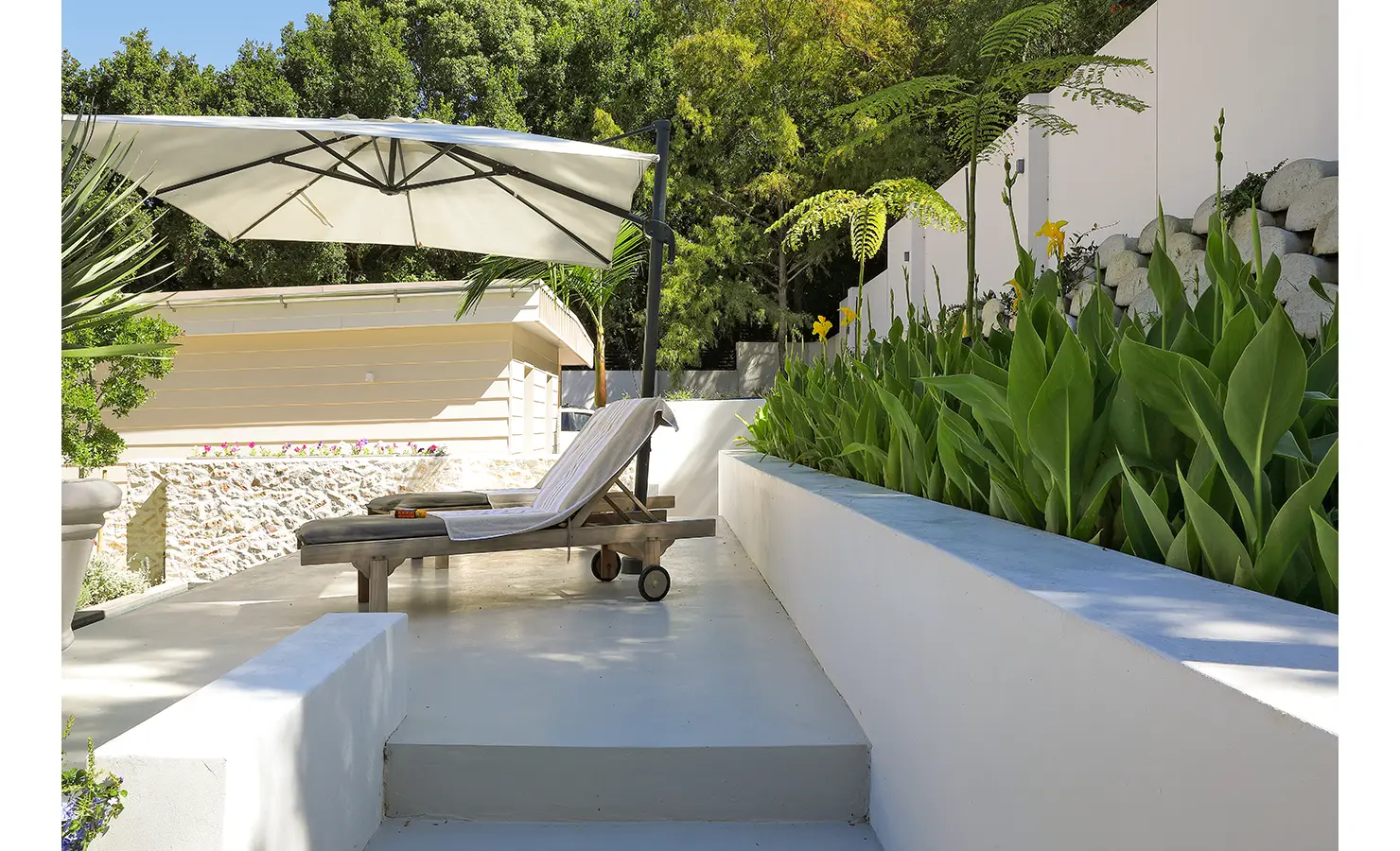 Perfect Hideaways: A serene terrace garden with white concrete walls, tropical plants, and a classic wooden lounger beneath a large sun umbrella. Bel Ombre Hills, Constantia.