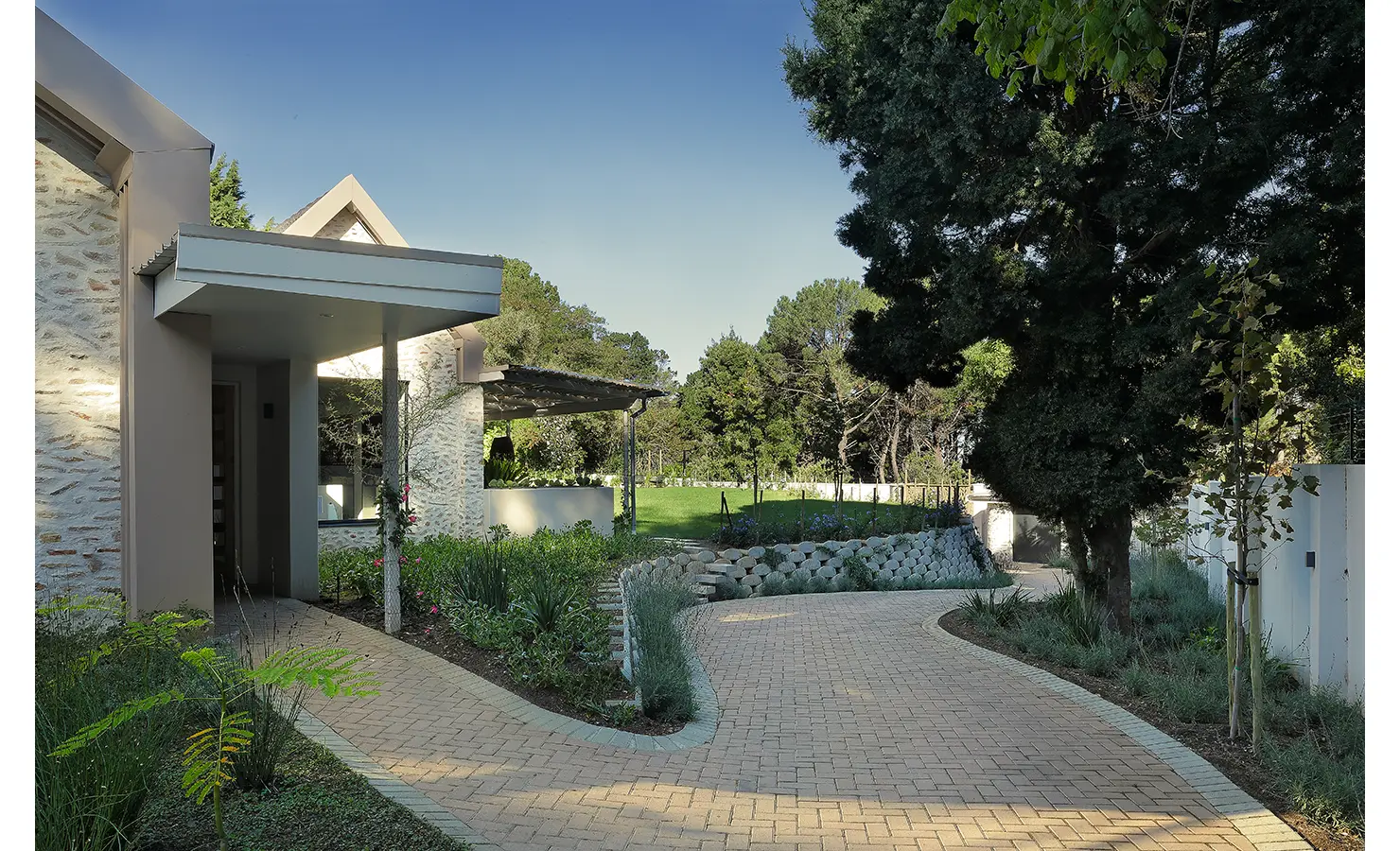 Perfect Hideaways: A curving paved driveway leads through manicured gardens to the front entrance, framed by lush greenery and tall trees. Bel Ombre Hills, Constantia.