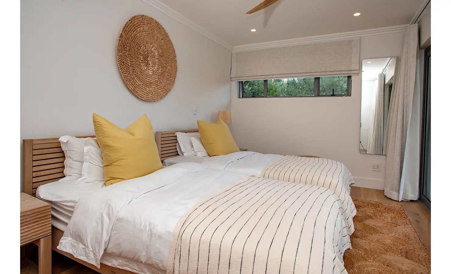 Perfect Hideaways: A cheerful twin bedroom styled in soft neutrals and mustard accents features slatted wooden beds, a woven wall hanging, and warm ambient lighting. River Run, Plettenberg Bay.