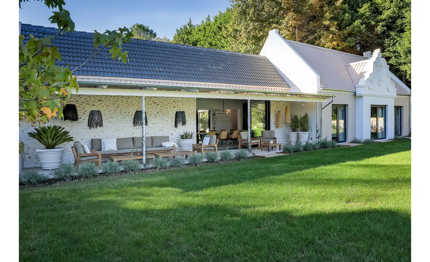 Perfect Hideaways: The exterior of a Cape Dutch-style home with a long shaded veranda, garden furniture, and vibrant lawn set against forested surroundings. Bel Ombre Hills, Constantia.