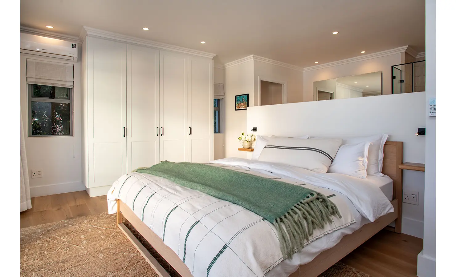 Perfect Hideaways: A serene bedroom with soft lighting, built-in white wardrobes, and a bed dressed in layered neutrals and a green throw, offers a tranquil retreat with elegant simplicity. River Run, Plettenberg Bay.