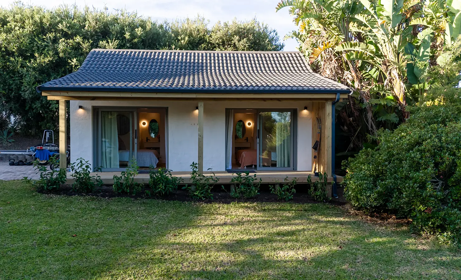 Perfect Hideaways: A charming garden cottage with a tiled roof and twin sliding doors reveals two warmly lit bedrooms nestled amidst lush greenery and tropical foliage. River Run, Plettenberg Bay.