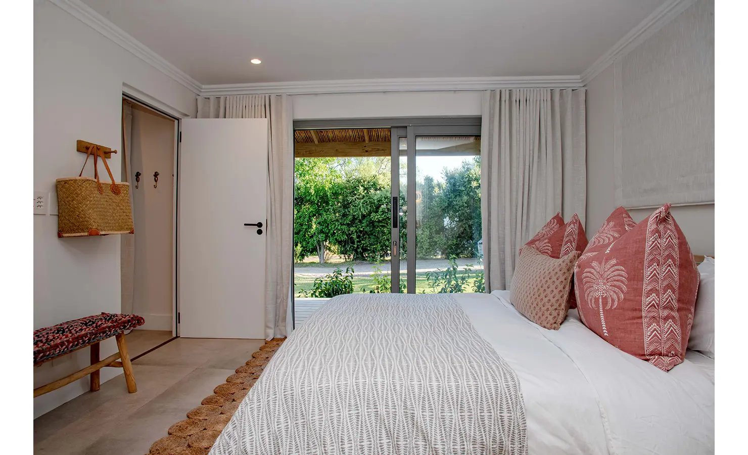 Perfect Hideaways: A cosy garden-facing bedroom with blush-toned cushions and textured bedding opens to verdant views through sliding doors, blending comfort and calm. River Run, Plettenberg Bay.