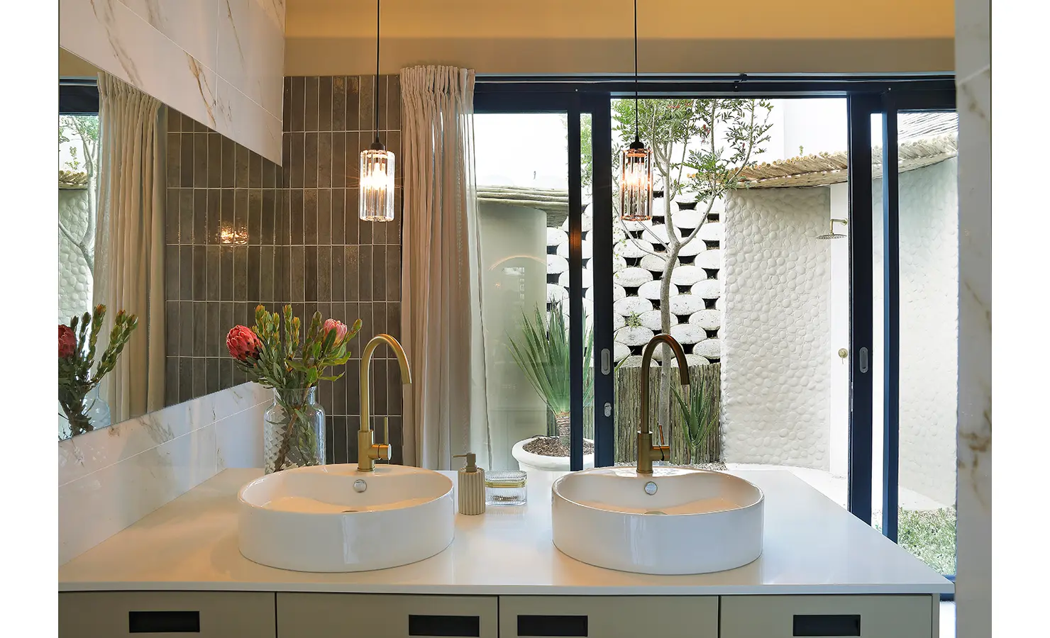 Perfect Hideaways: A chic bathroom interior with twin basins, brass fittings, and sliding glass doors that open to a secluded outdoor shower. Bel Ombre Hills, Constantia.