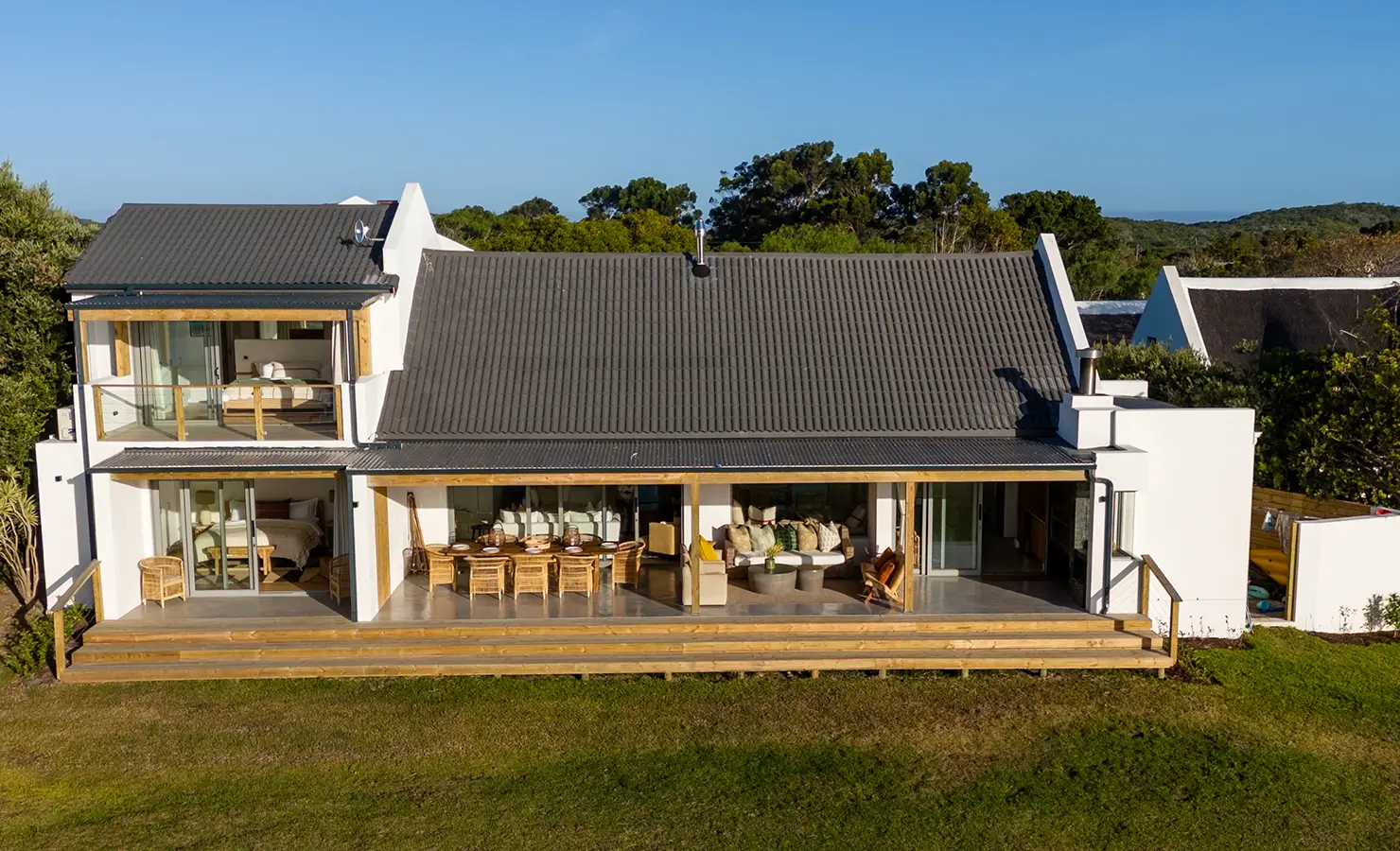 Perfect Hideaways: A wide-angle view of the house shows its expansive covered veranda, upper balcony, and seamless indoor-outdoor layout, all set against a lush natural backdrop. River Run, Plettenberg Bay.