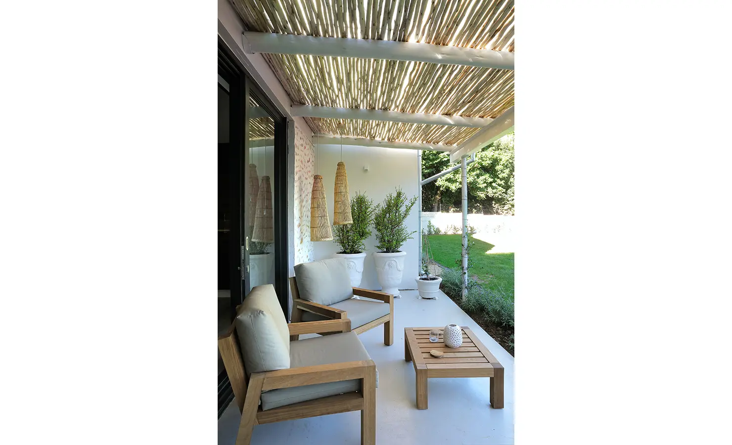 Perfect Hideaways: A peaceful veranda with wooden armchairs, soft grey cushions, and a low table under a reed pergola, overlooking manicured lawns. Bel Ombre Hills, Constantia.