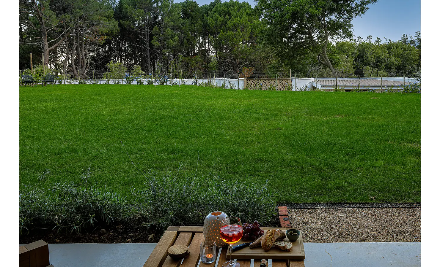 32 Perfect Hideaways: A serene view across a vibrant green lawn from a shaded terrace, with a table set for sunset drinks and nibbles. Bel Ombre Hills, Constantia.