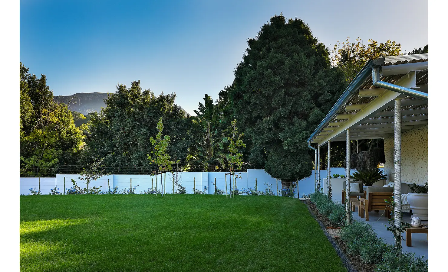 Perfect Hideaways: A sweeping lawn bordered by trees and a covered veranda, with mountain peaks visible in the distance under a clear blue sky. Bel Ombre Hills, Constantia.