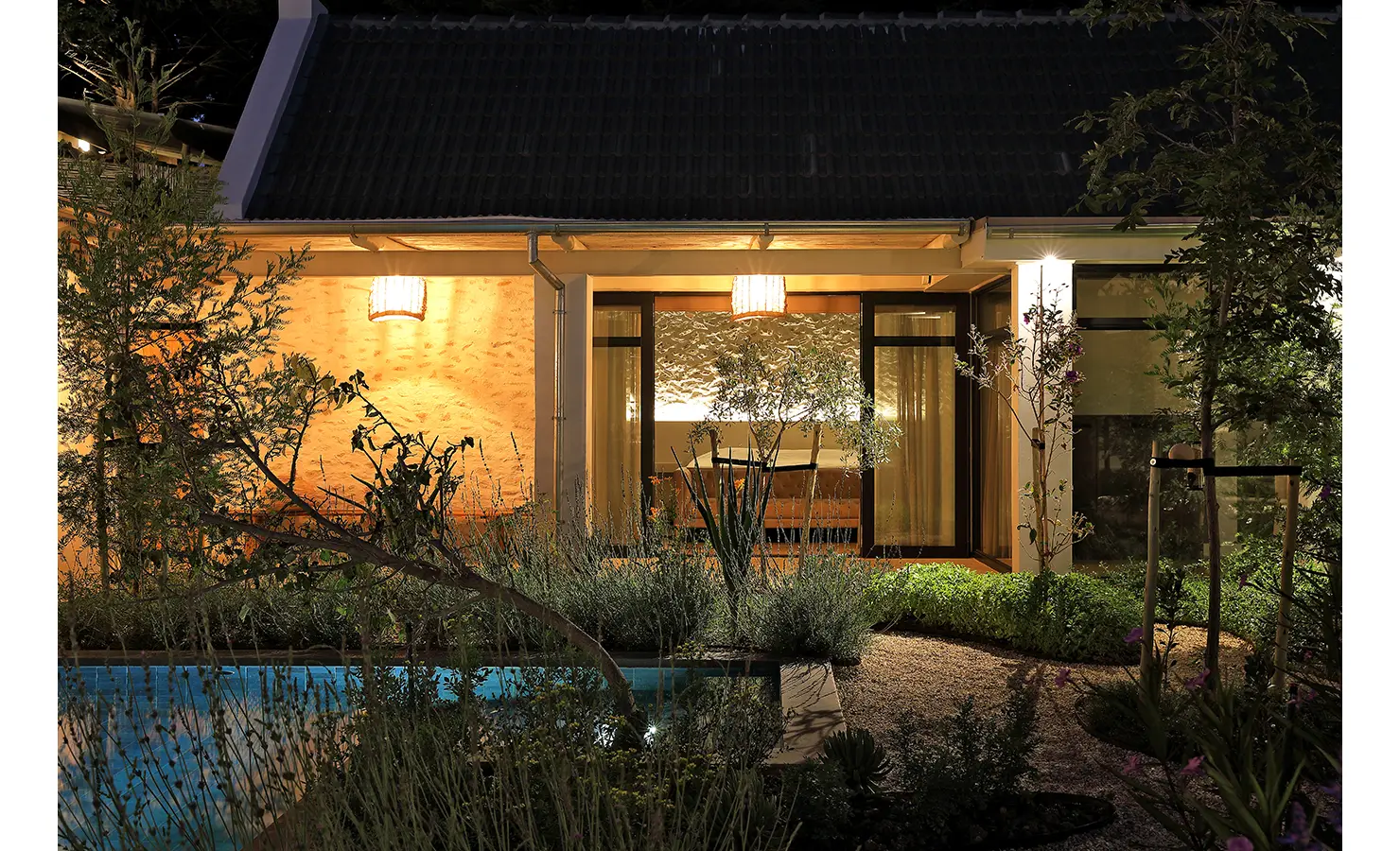 Perfect Hideaways: A warmly lit garden façade at night, with soft light glowing from inside and lush landscaping around the pool’s edge. Bel Ombre Hills, Constantia.