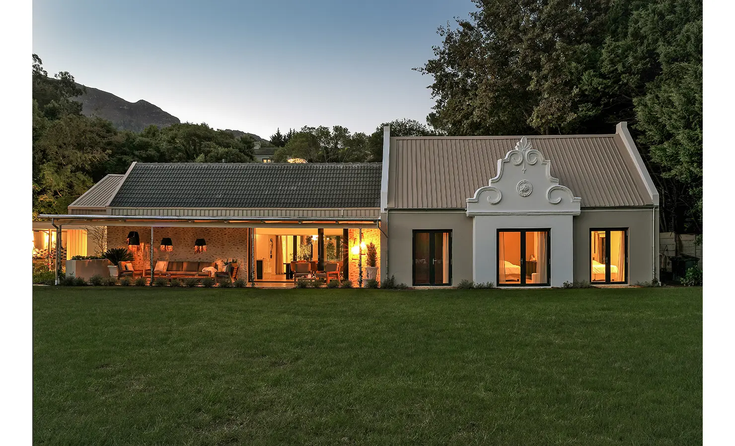 Perfect Hideaways: The full exterior of the Cape Dutch-style home at dusk, warmly lit from within and framed by a large lawn and forest backdrop. Bel Ombre Hills, Constantia.