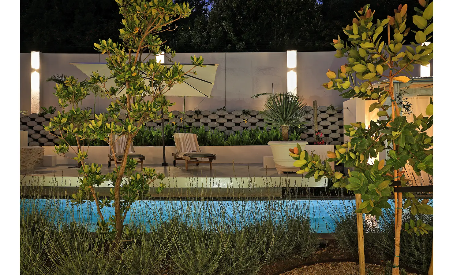 Perfect Hideaways: A nighttime view of the pool and garden loungers under glowing wall sconces, framed by lush tropical foliage and a curved wall of stones. Bel Ombre Hills, Constantia.