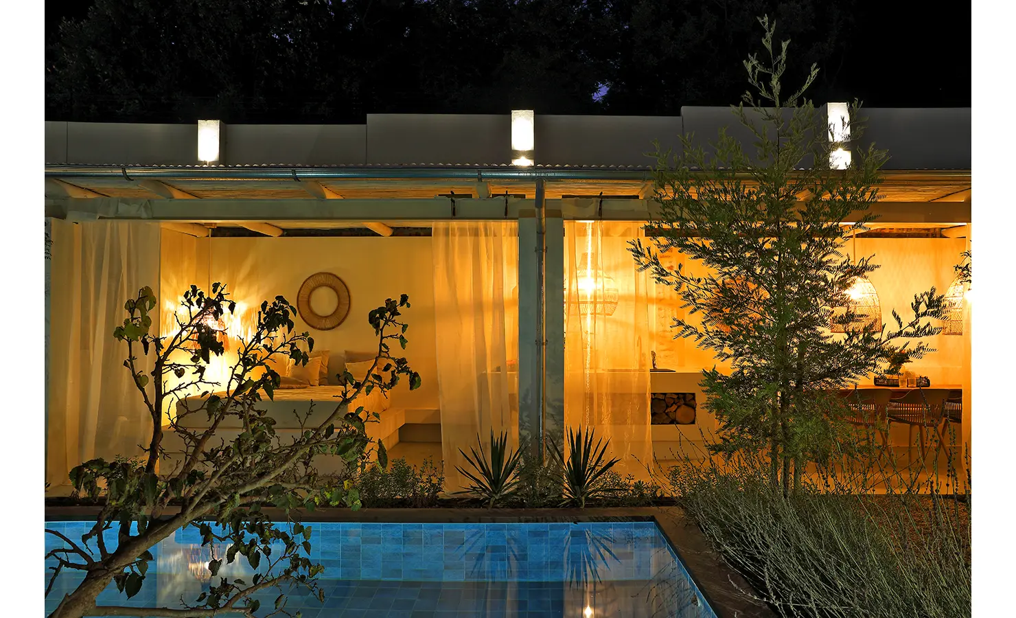 Perfect Hideaways: A glowing poolside pavilion at night, softly lit through sheer curtains with views into the outdoor kitchen and lounge. Bel Ombre Hills, Constantia.