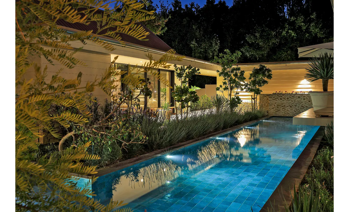Perfect Hideaways: A beautifully lit pool at twilight reflecting soft garden lighting, framed by planting and the home’s pale façade. Bel Ombre Hills, Constantia.