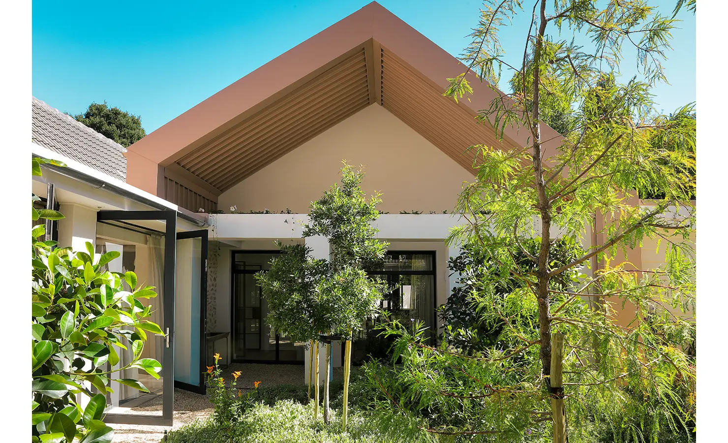 Perfect Hideaways: A modern architectural façade with a striking gabled roof rises behind a lush, sunlit courtyard filled with greenery and young trees. Bel Ombre Hills, Constantia.