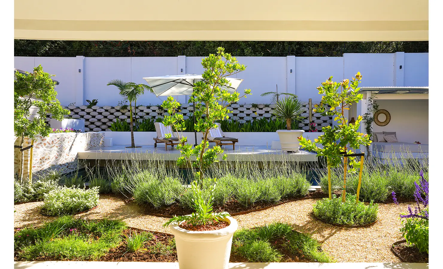 Perfect Hideaways: A tranquil garden scene with manicured planting beds, lavender, and young trees, facing a pool terrace with sun loungers and a shaded pavilion. Bel Ombre Hills, Constantia.