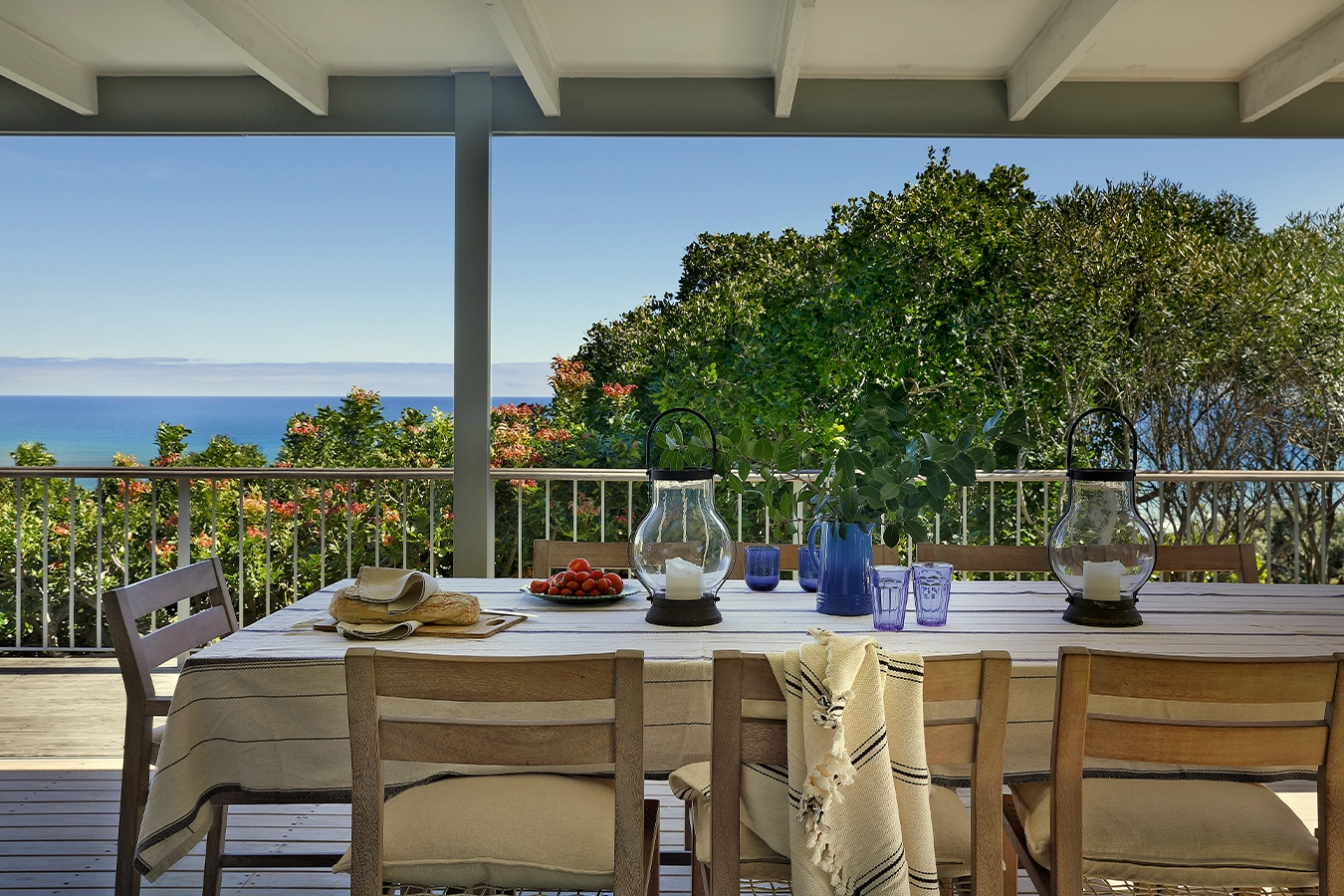 Perfect Hideaways: Shaded verandah dining table with a striped cloth, storm lanterns, blue glassware, bread and tomatoes, framed by treetops and a calm turquoise ocean on the horizon — relaxed alfresco living, Amongst The Trees, Scarborough.