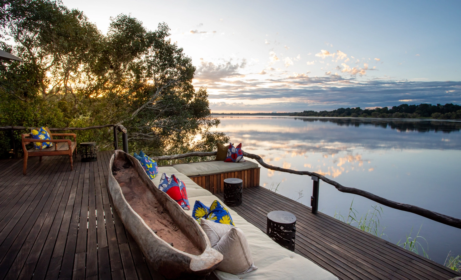 Perfect Hideaways: riverfront timber deck with cushioned built-in daybeds, colourful scatter cushions and a rustic dugout canoe centrepiece, overlooking still water and a pastel sunset sky — Mukwa River Lodge, Livingstone