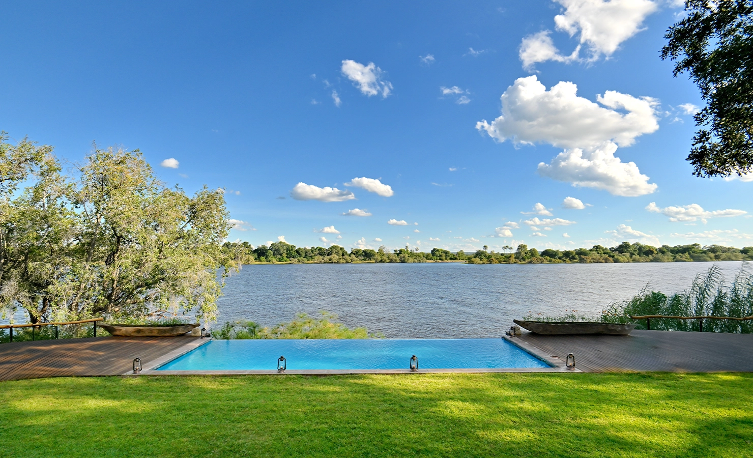 Perfect Hideaways: Infinity pool overlooking a wide, tranquil river with a lush green lawn in the foreground and a bright blue sky dotted with fluffy clouds in the background, Tanyati Villa, Livingstone