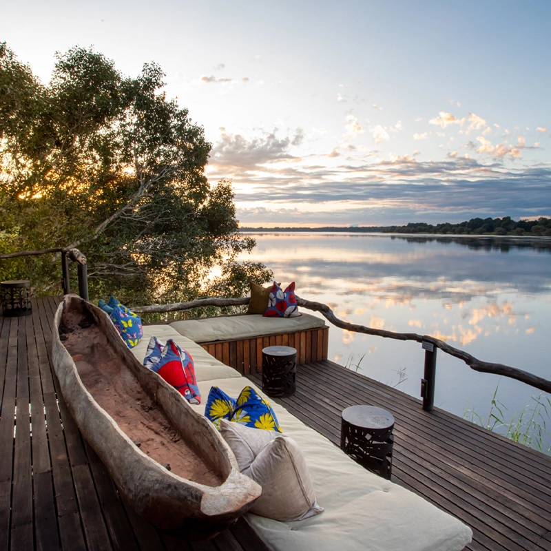 Perfect Hideaways: Expansive wooden deck with cushioned seating, vibrant patterned pillows, and a traditional carved canoe overlooking the tranquil Zambezi River at sunset, Mukwa River Lodge, Livingstone.