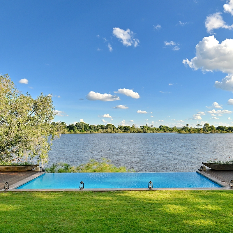Perfect Hideaways: Infinity pool overlooking a wide, tranquil river with a lush green lawn in the foreground and a bright blue sky dotted with fluffy clouds in the background, Tanyati Villa, Livingstone