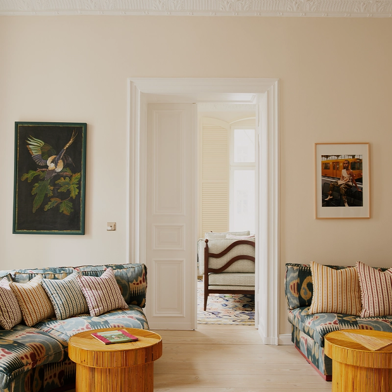 Perfect Hideaways: A welcoming salon adorned with a patterned curved sofa lined with striped cushions, a red pleated floor lamp, and framed artworks including a bird illustration and a portrait; the space flows into a bright bedroom through double doors, creating a sense of openness and harmony. The Artists Nest, Kollwitzkiez
