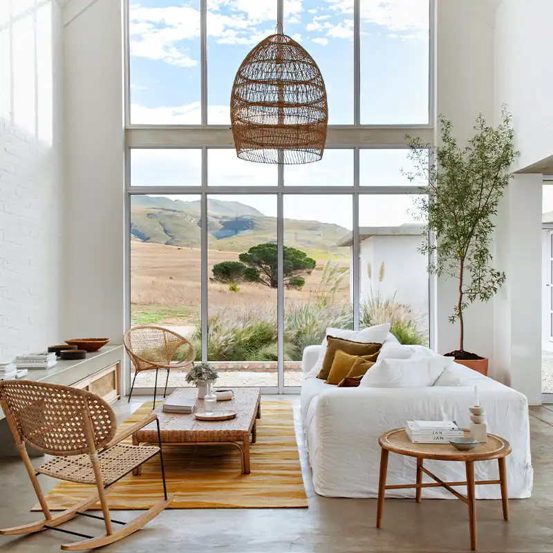Perfect Hideaways: Spacious living room with large windows showcasing a scenic landscape. Features a cozy white sofa, woven furniture, and a warm, airy atmosphere. Shoot Locations.