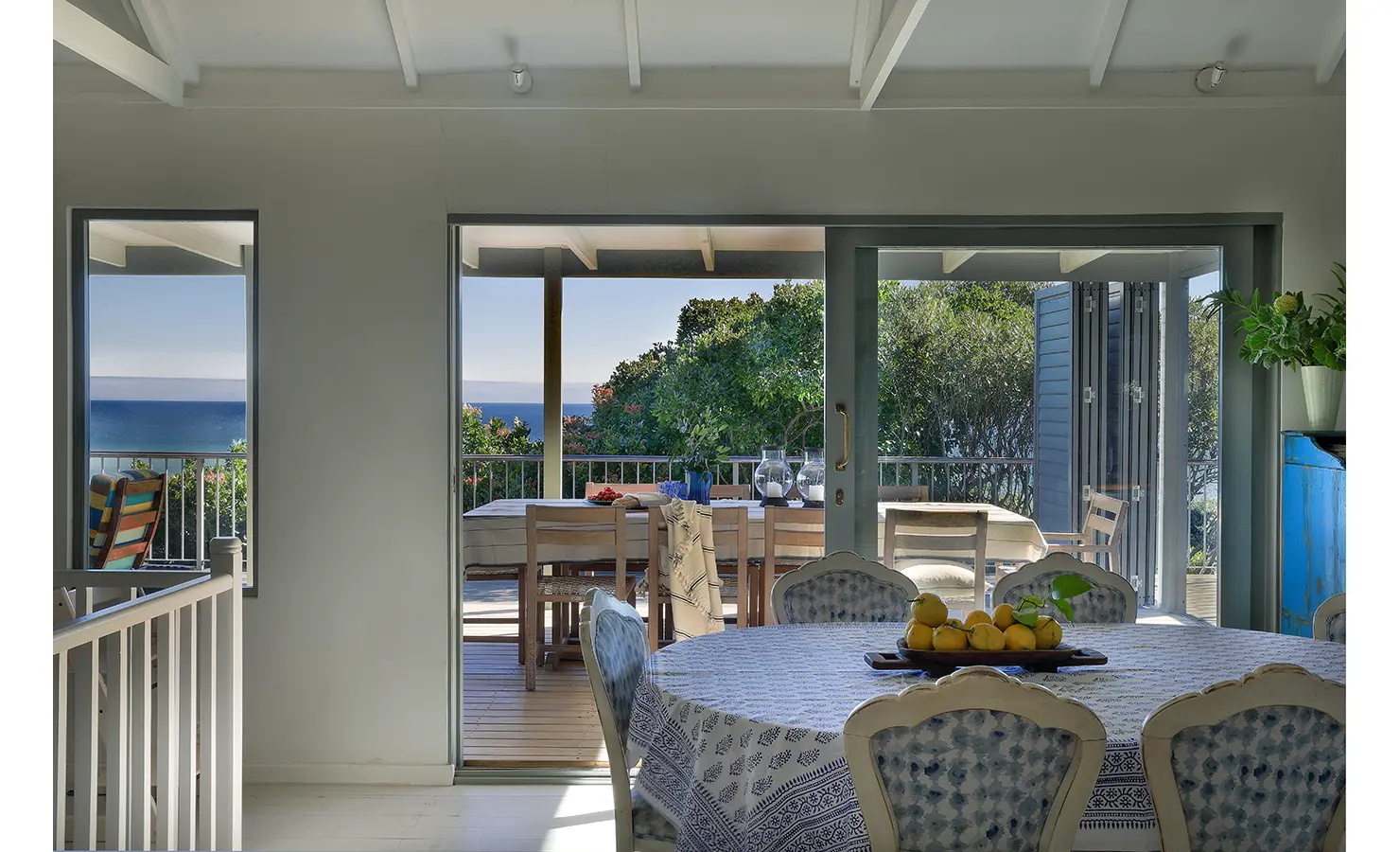 Perfect Hideaways: Blue-and-white dining table with a bowl of lemons, doors opening to the shaded deck framed by treetops and an ocean horizon — easy indoor-outdoor flow, Amongst The Trees, Scarborough.