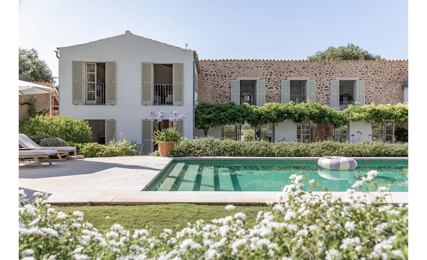 Perfect Hideaways: A traditional Mallorcan farmhouse façade of stone and whitewashed walls, lined with soft green shutters and framed by lush wisteria, overlooking a glistening swimming pool with sun loungers and flowering borders. Finca Es Rafal, Alaró