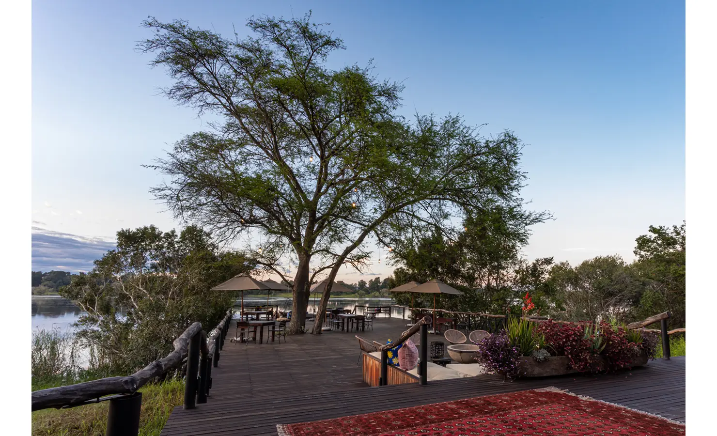 Perfect Hideaways: expansive riverfront deck beneath a mature tree with dining tables, parasols, relaxed seating and lush planters at the water’s edge — Mukwa River Lodge, Livingstone