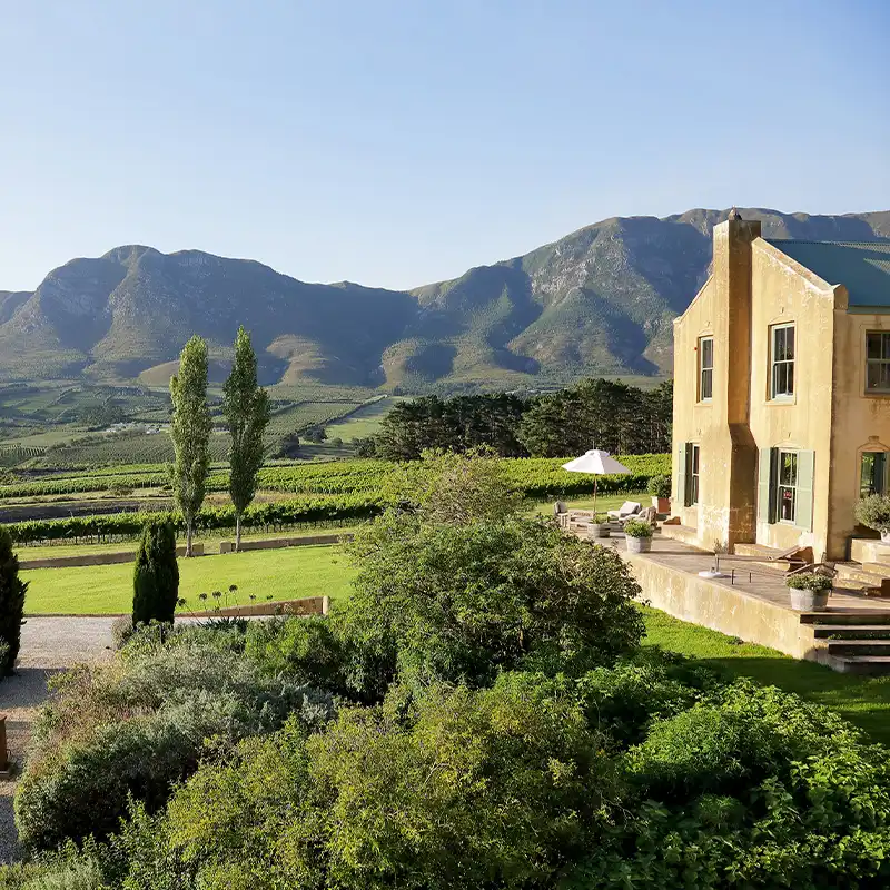 Perfect Hideaways: A sunlit countryside villa overlooks lush vineyards and rolling hills, framed by distant mountains under a clear blue sky. The scene is serene and expansive. Shoot Locations.