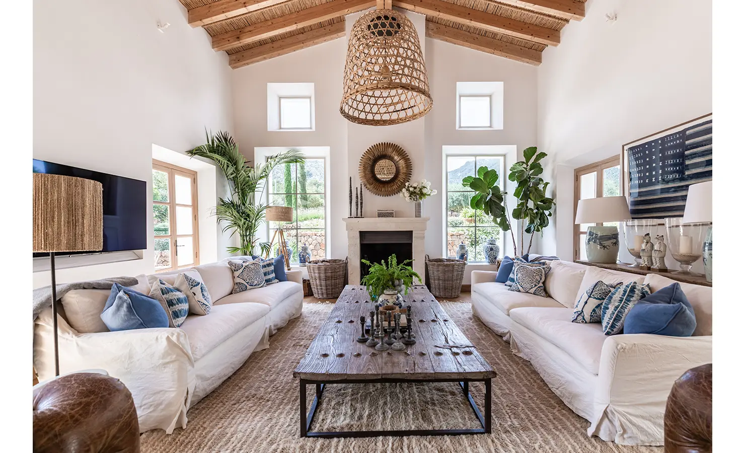 Perfect Hideaways: A light-filled sitting room with high ceilings and exposed timber beams, styled with soft white sofas, botanical touches, and indigo cushions, centred around a rustic fireplace and vintage coffee table. Finca Es Rafal, Alaró