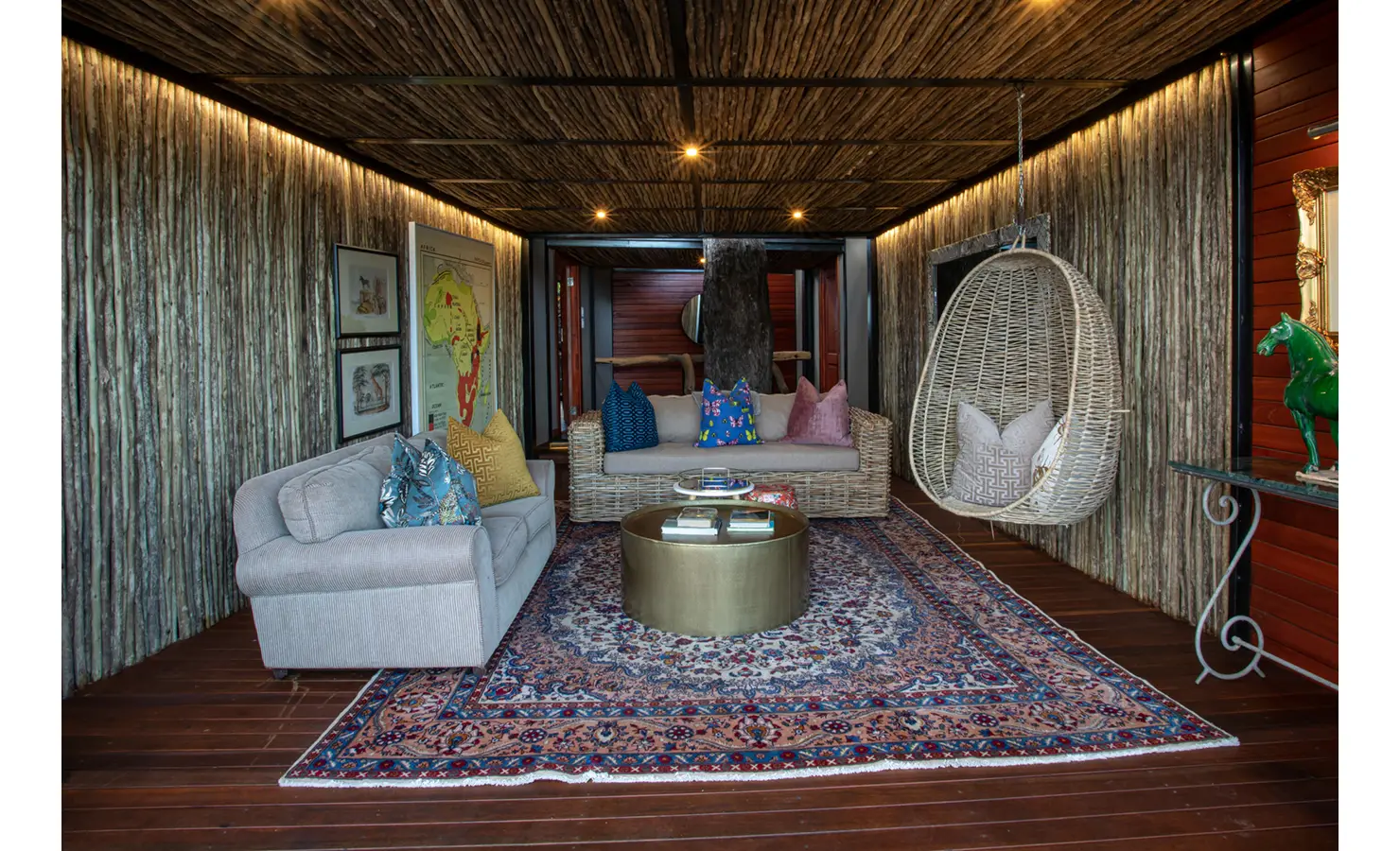 Perfect Hideaways: cosy lounge with rattan sofa, upholstered couch and a hanging wicker egg chair on a Persian rug, rustic stick-clad walls and a central tree-trunk feature — Mukwa River Lodge, Livingstone