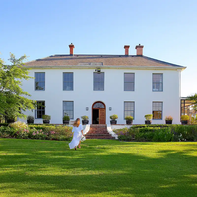 Perfect Hideaways: A woman in a white dress walks toward a large, elegant white house with a symmetrical facade, set in a lush green garden under a clear blue sky. Shoot Locations.