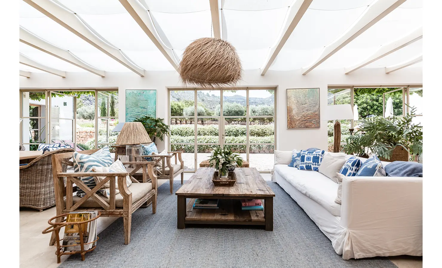Perfect Hideaways: A breezy sunroom flooded with natural light, featuring relaxed wicker furniture, nautical blue and white accents, and views through floor-to-ceiling glass doors to manicured gardens beyond. Finca Es Rafal, Alaró