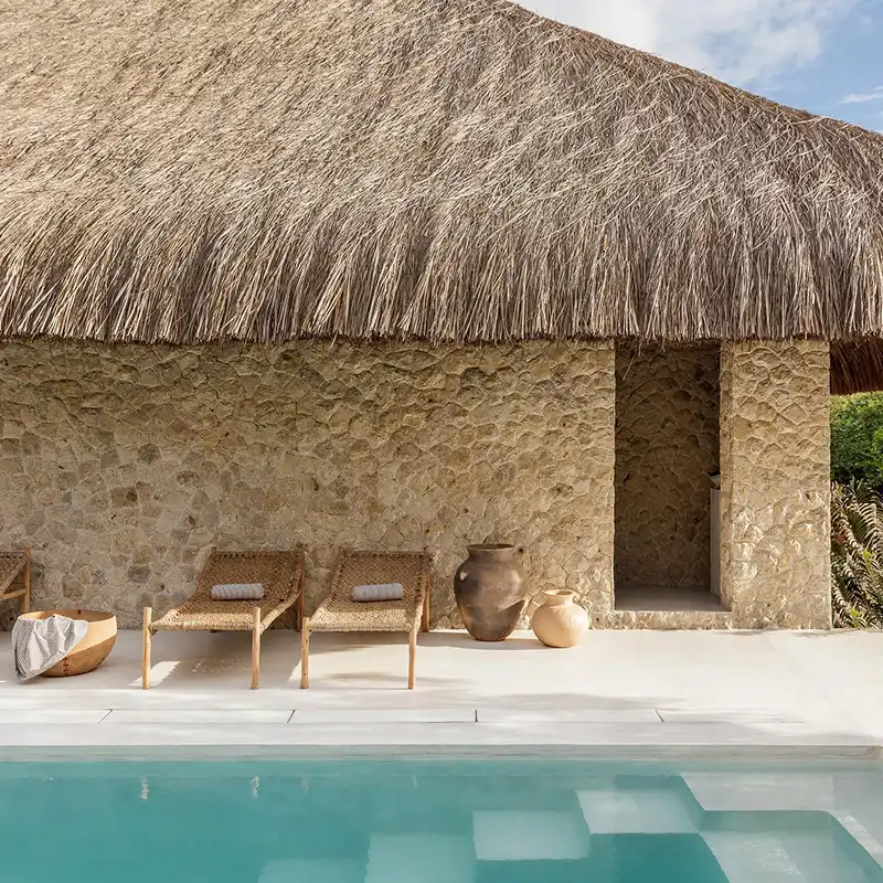 Perfect Hideaways: Rustic stone-walled hut with a thatched roof by a pool. Two woven chairs, a rolled towel, and clay pots create a relaxing, tropical vibe. Shoot Locations.