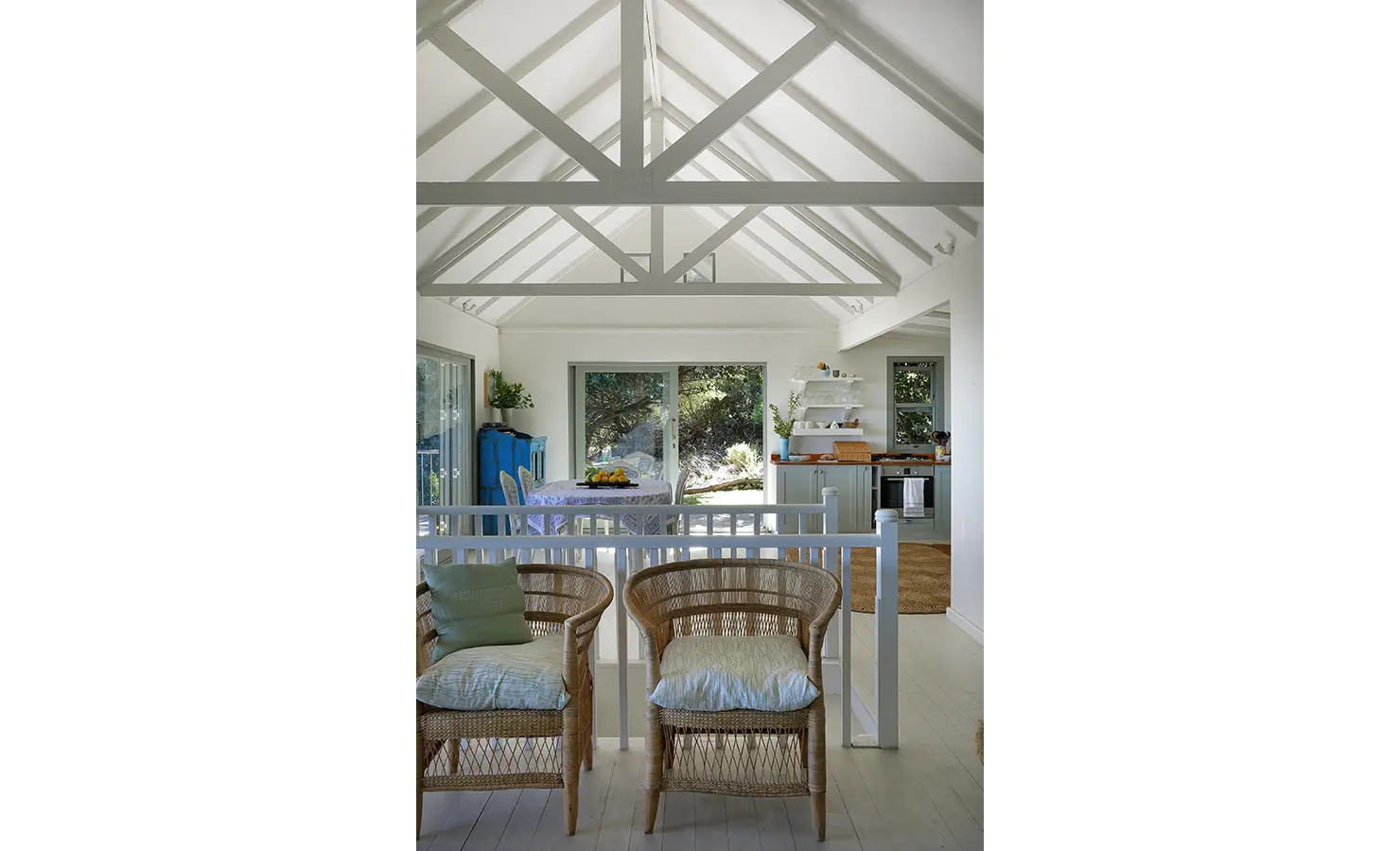 Perfect Hideaways: Double-volume ceiling with exposed trusses above airy open-plan living; rattan armchairs lead through to kitchen and dining, Amongst The Trees, Scarborough.