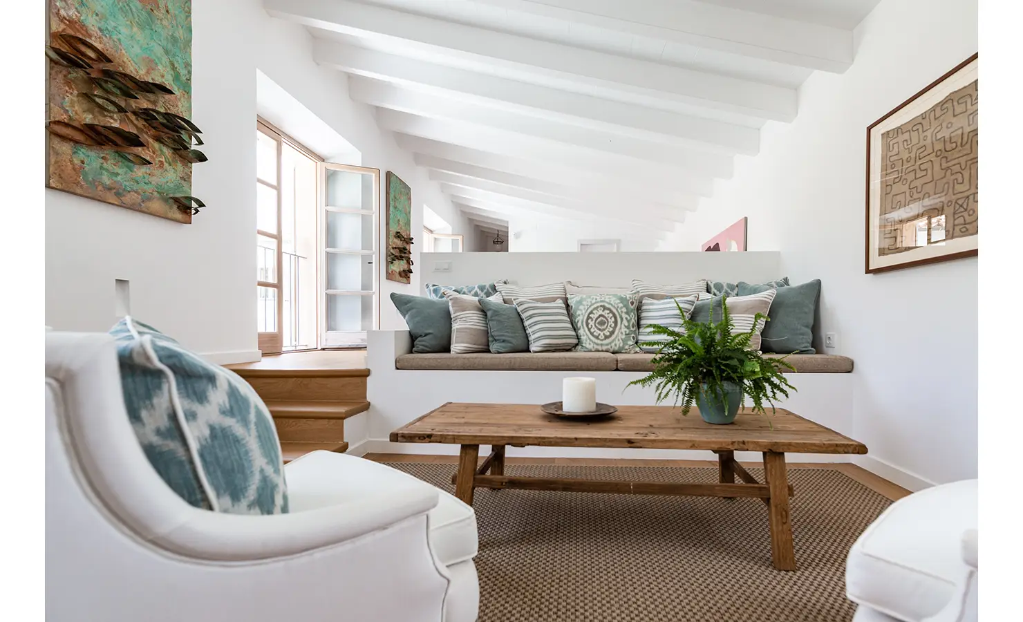 Perfect Hideaways: A cosy mezzanine-style lounge nook with built-in bench seating adorned in sage and patterned cushions, set beneath slanted white beams and flanked by textural art and soft natural light. Finca Es Rafal, Alaró