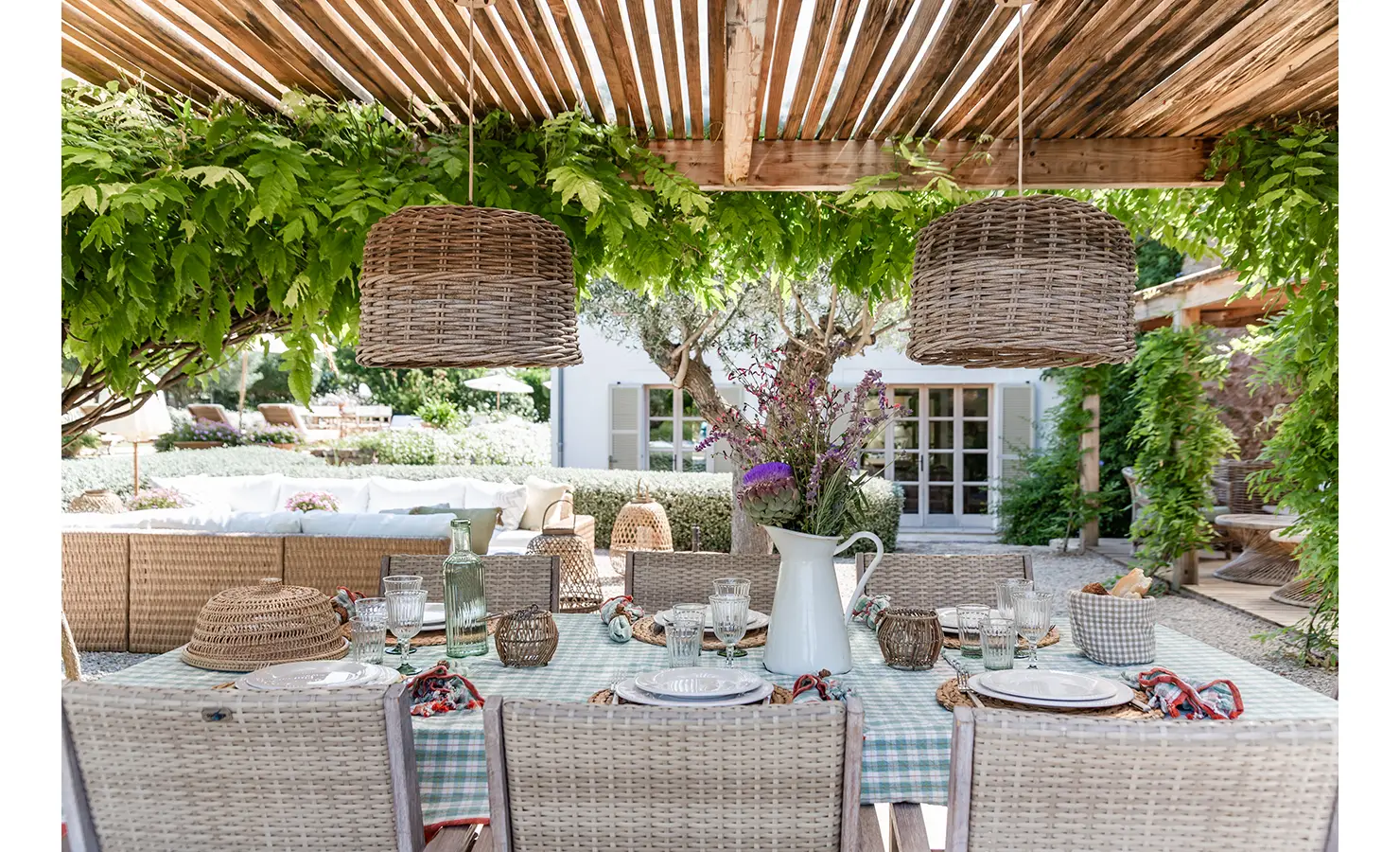 Perfect Hideaways: An enchanting outdoor dining space shaded by a timber pergola and lush vines, styled with a gingham tablecloth, woven chairs, and layered table settings under rustic basket pendant lights. Finca Es Rafal, Alaró