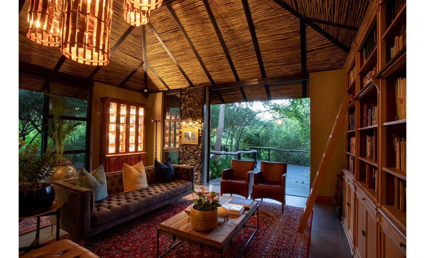 Perfect Hideaways: library lounge with tufted sofa, leather armchairs, timber bookshelves with ladder and a view to the deck and trees — Mukwa River Lodge, Livingstone