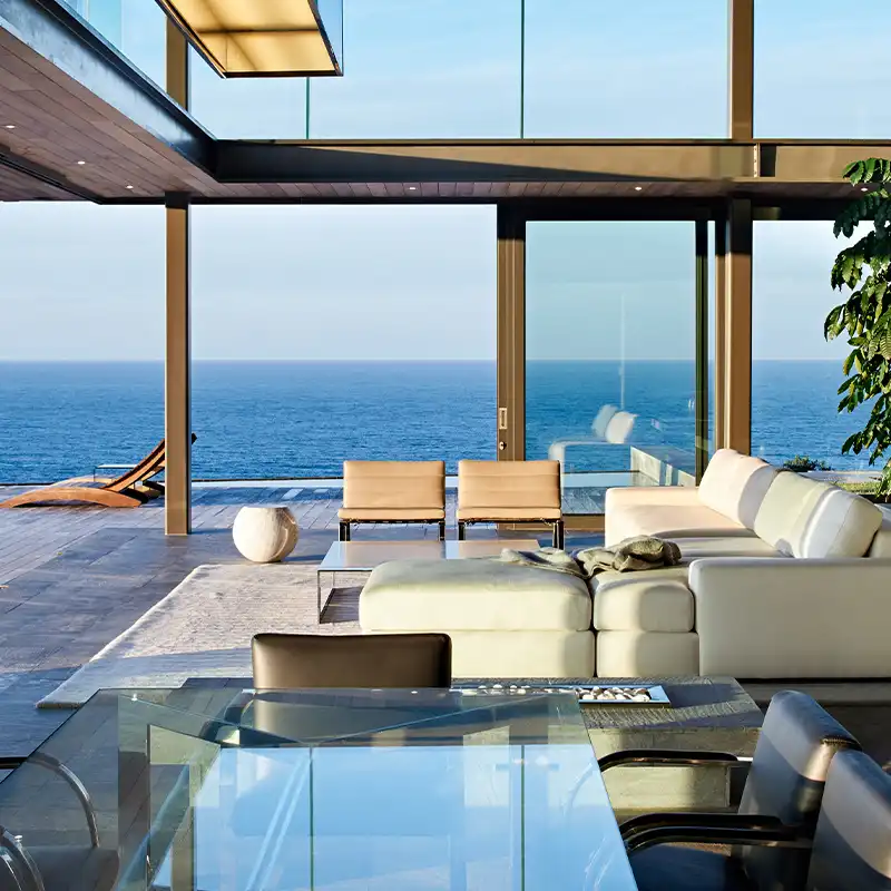 Perfect Hideaways: Modern living room with glass walls overlooking the ocean. Features a white couch, beige chairs, glass table, and sunny, serene atmosphere. Shoot Locations.