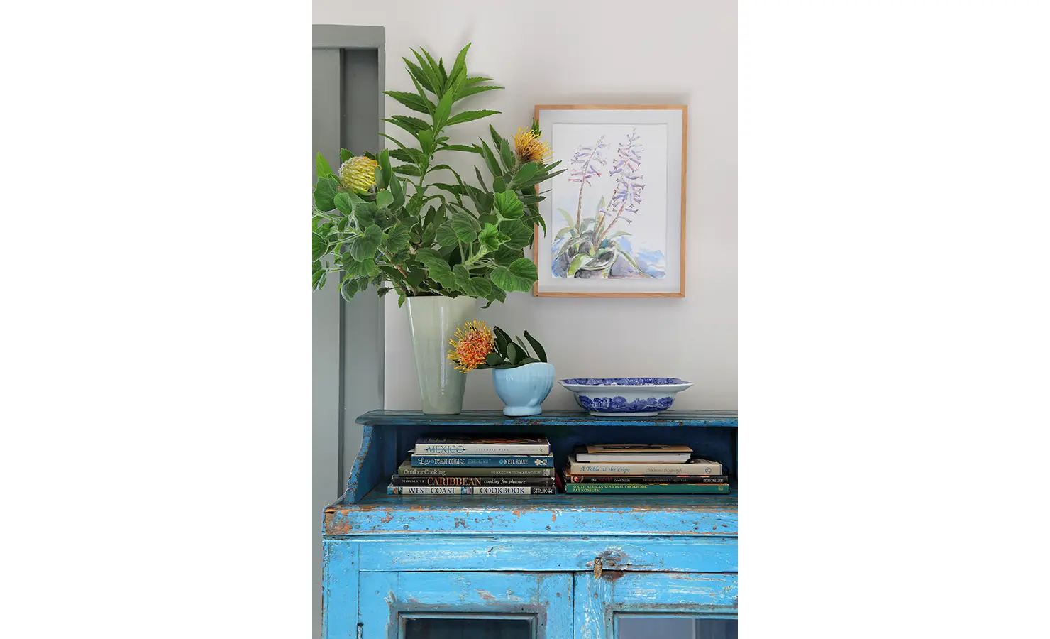 Perfect Hideaways: Styled vignette — protea-like blooms and leafy greens with ceramics and a botanical watercolour atop a distressed azure cabinet, Amongst The Trees, Scarborough.