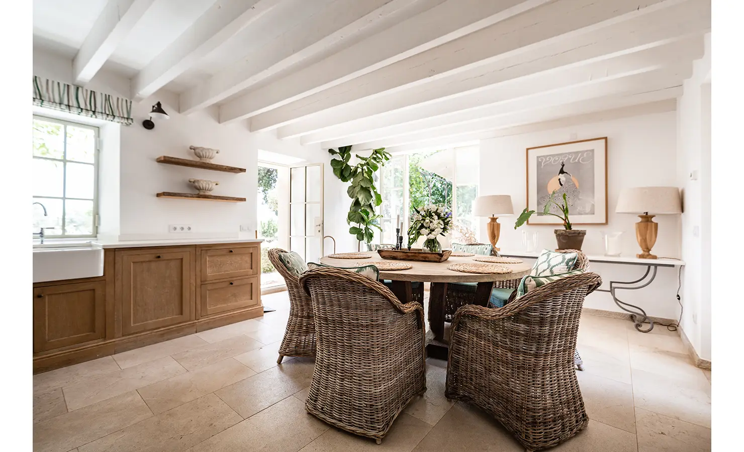 Perfect Hideaways: A charming country-style kitchen and dining area with wicker armchairs, a circular wooden table, and botanical accents bathed in soft light beneath white ceiling beams. Finca Es Rafal, Alaró