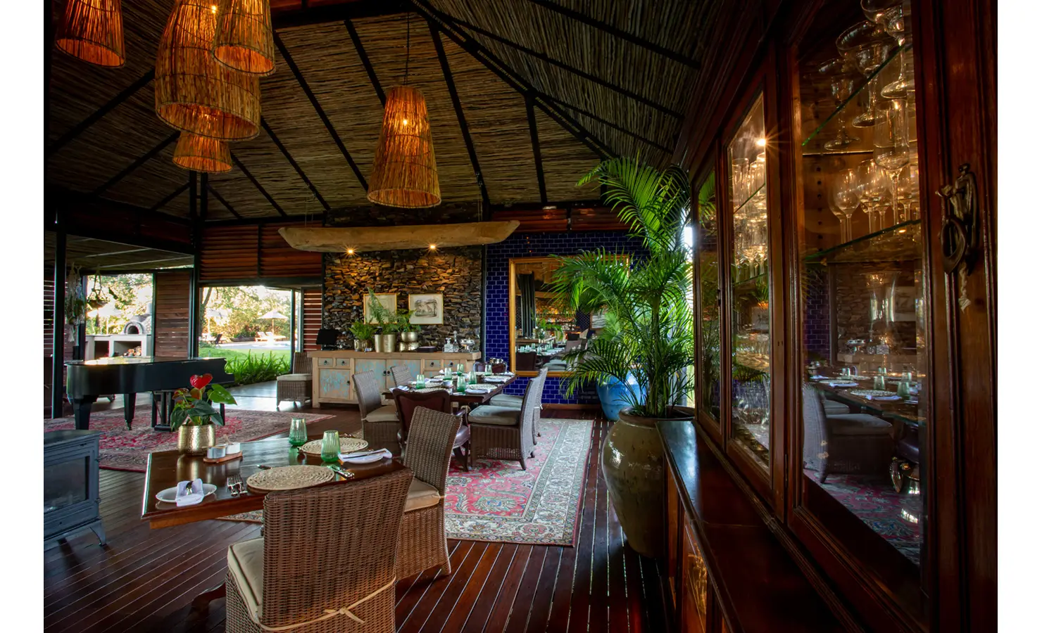 Perfect Hideaways: airy dining verandah with piano, cane chairs, potted palms and glass display cabinet, set beneath sculptural woven pendants — Mukwa River Lodge, Livingstone