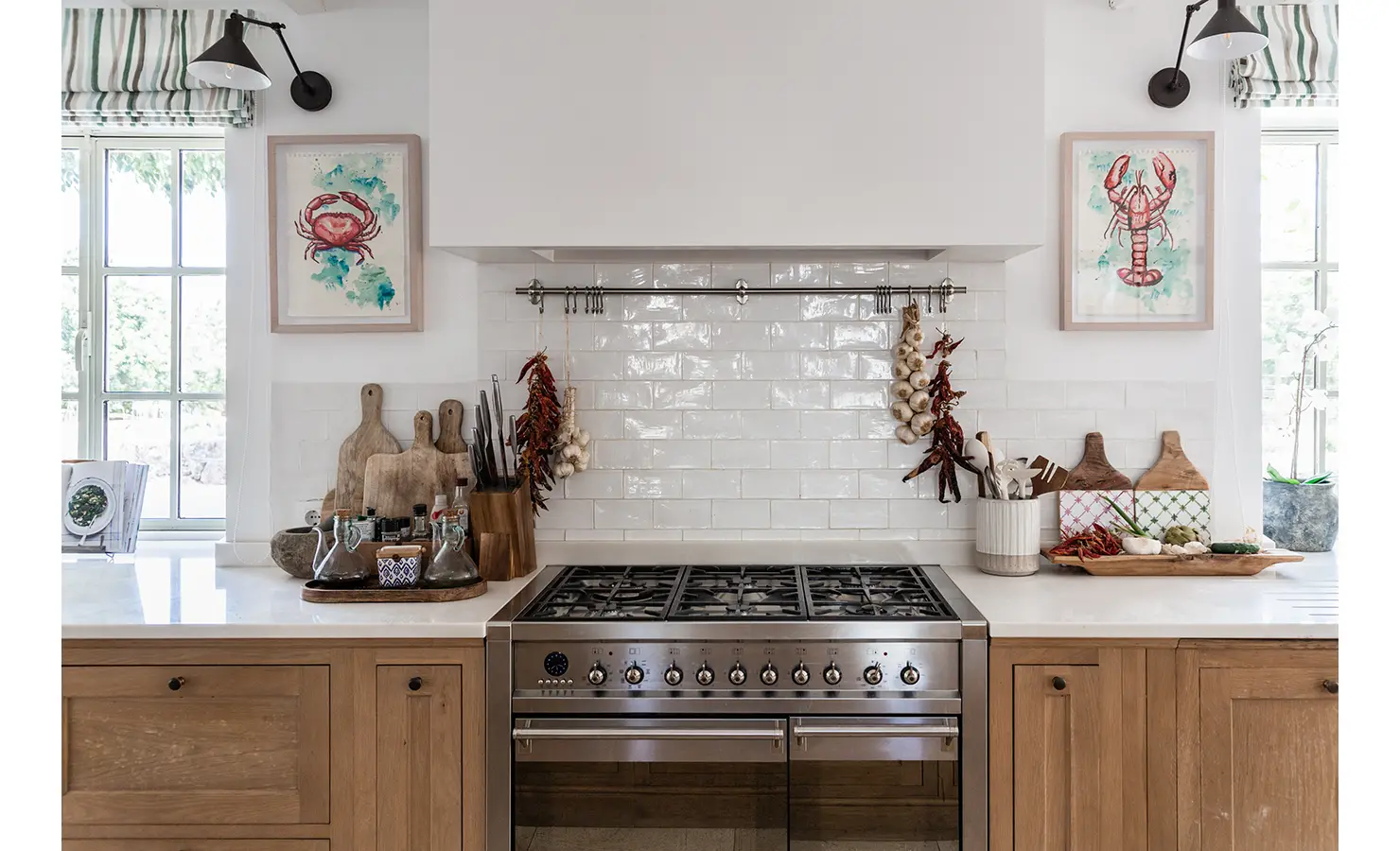 Perfect Hideaways: A warm and welcoming kitchen vignette with a professional gas stove framed by white subway tiles, hanging garlic and chillies, and rustic chopping boards beneath playful crustacean artwork. Finca Es Rafal, Alaró